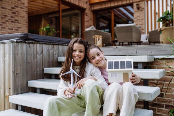 Sisters holding model of house with solar panels on roof and wind turbine model. Alternative, renewable, green energy and sustainable lifestyle for future generations concept.