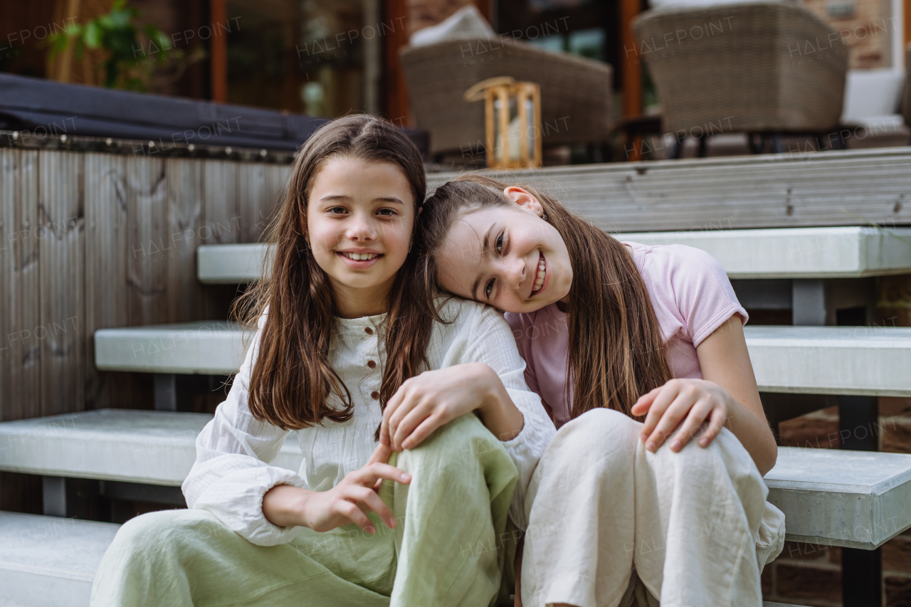 Two sisters sitting on front porch stairs in front of house, embracing. Sisterly love and siblings relationship concept.