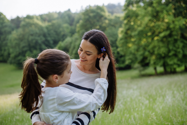 Beautiful mother with daughter, picking flowers, putting in moms, hair, sitting in the grass at meadow. Concept of Mother's Day and maternal love.