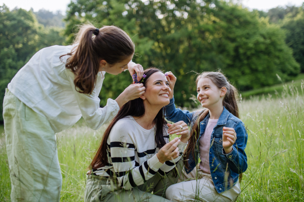 Beautiful mother with daughters, picking flowers, putting in moms, hair, sitting in the grass at meadow. Concept of Mother's Day and maternal love.