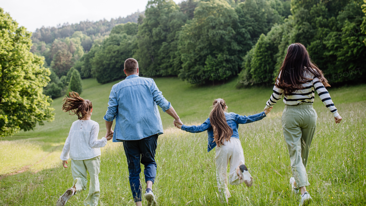 Rear view of beautiful young family walking, running through grass at meadow, enjoying together time.