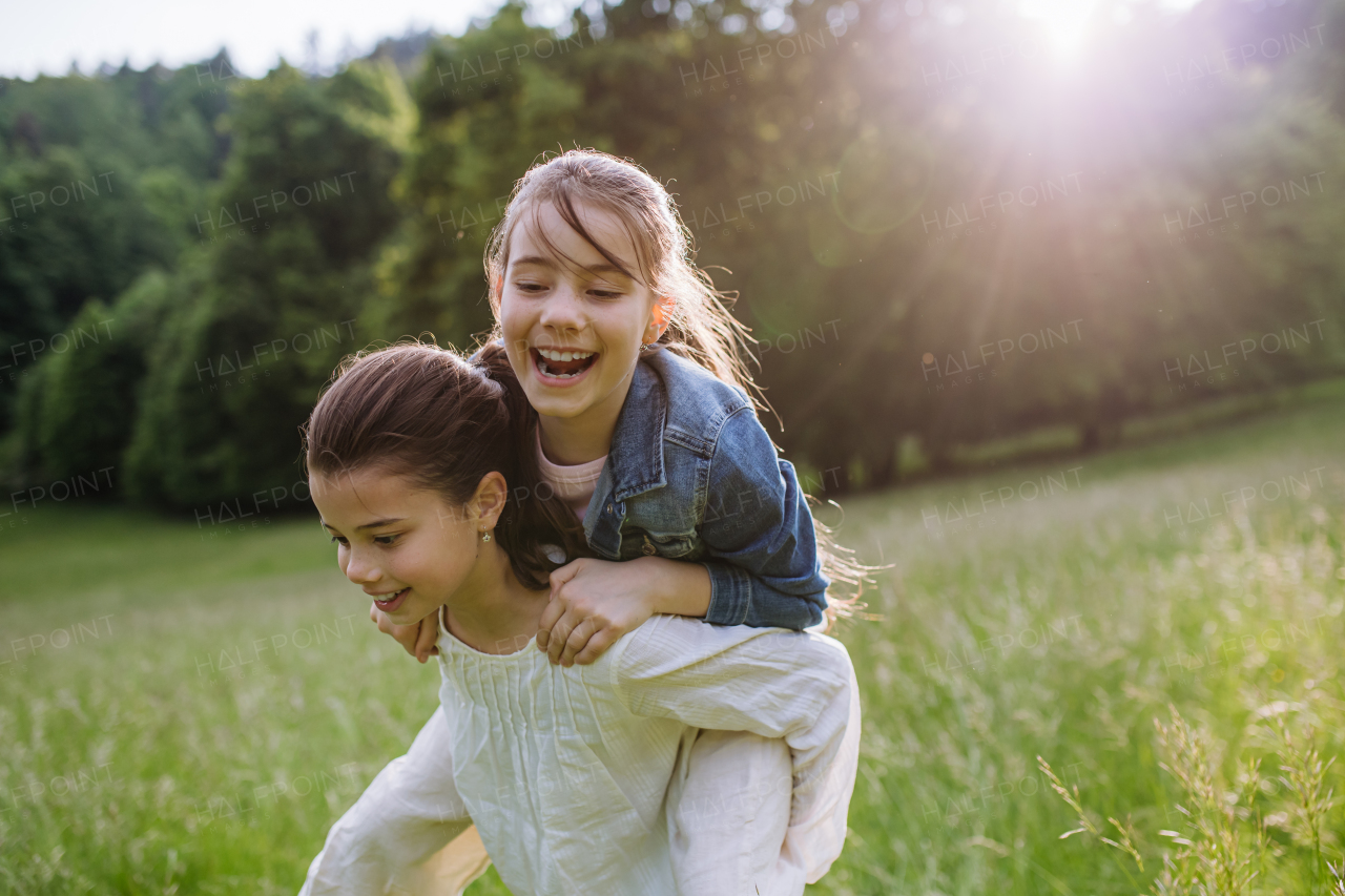 Two sisters playing at meadow in tall grass, having fun, piggybacking each other. Sisterly love and siblings relationship concept.