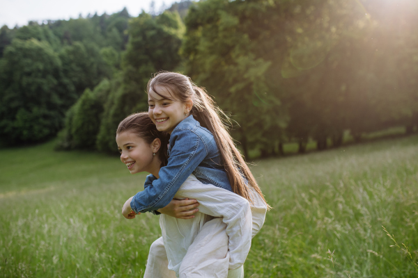 Two sisters playing at meadow in tall grass, having fun, piggybacking each other. Sisterly love and siblings relationship concept.