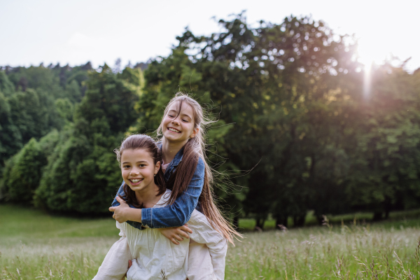 Two sisters playing at meadow in tall grass, having fun, piggybacking each other. Sisterly love and siblings relationship concept.