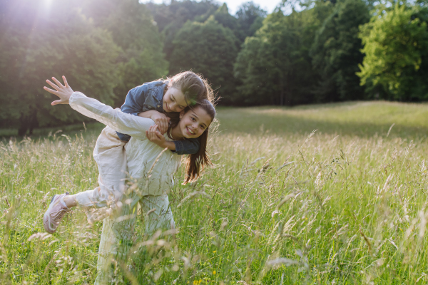 Two sisters playing at meadow in tall grass, having fun, piggybacking each other. Sisterly love and siblings relationship concept.