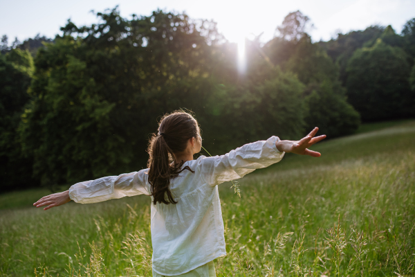 Rear view of young girl standing on meadow with open arms, enjoying warm spring day and sun.