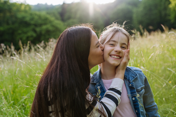 Beautiful mother with daughter, embracing and kissing on cheek, sitting in the grass at meadow. Concept of Mother's Day and maternal love.