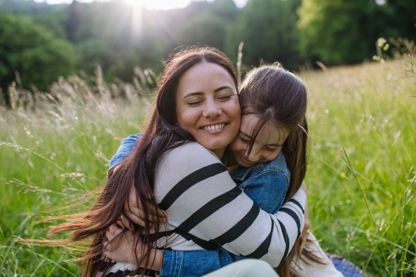 Beautiful mother with daughter, embracing or hugging, sitting in the grass at meadow. Concept of Mother's Day and maternal love.