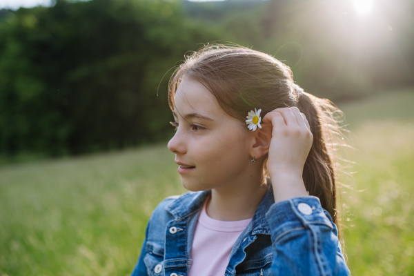 Portrait of beautiful young girl with flower behind the ear, standing on meadow, enjoying warm spring day.