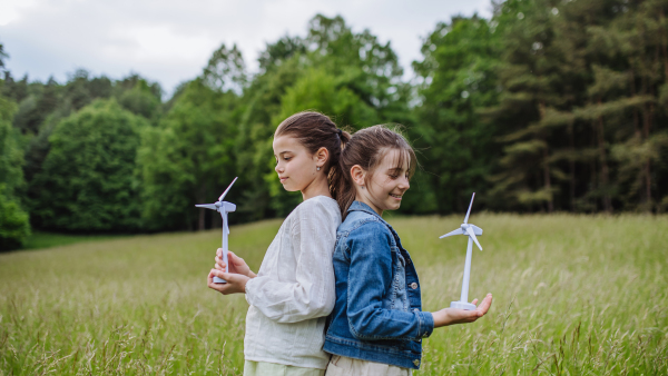 Two girls holding model wind turbine, standing on meadow, in nature. Alternative, renewable, green energy and sustainable lifestyle for future generations concept.