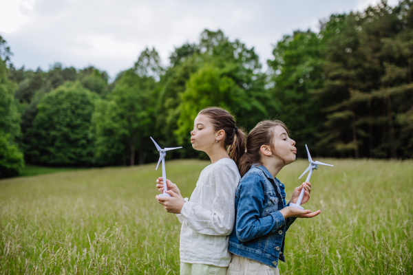 Two girls holding model wind turbine, standing on meadow, in nature. Alternative, renewable, green energy and sustainable lifestyle for future generations concept.