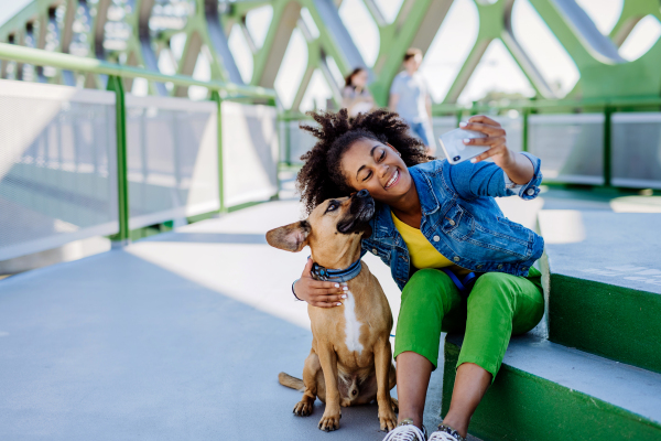 Multiracial girl sitting and resting with her dog outside in the bridge, taking selfie with him, spending leisure time together. Concept of everyday life with pet.