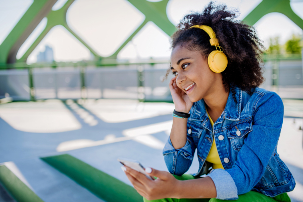 Multiracial teenage girl with a backpack, headphones and smartphone resting after school in city bridge, listening to music.