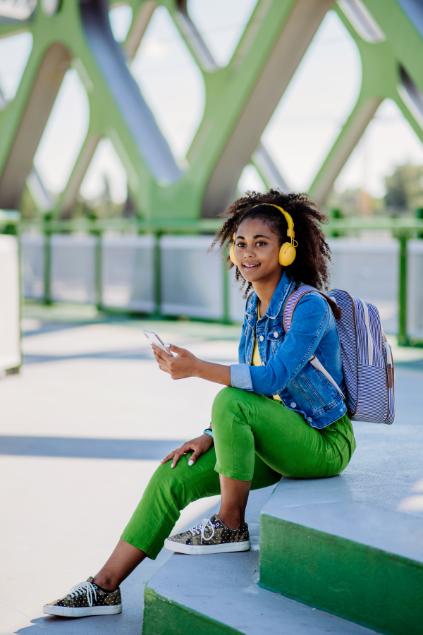 Multiracial teenage girl with a backpack, headphones and smartphone resting after school in city bridge, listening to music.