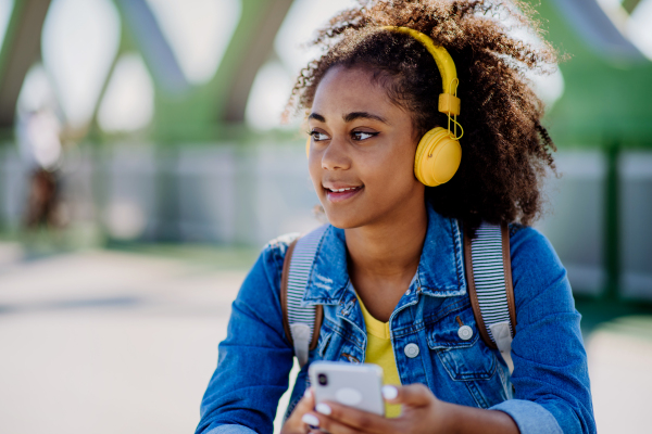 Multiracial teenage girl with a backpack, headphones and smartphone resting after school in city bridge, listening to music.