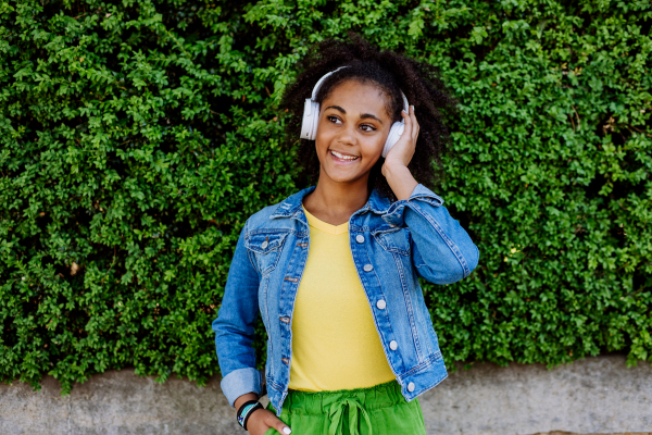 Multiracial girl enjoying music in the headphones, standing in front of green bush, portrait.