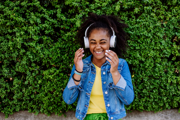 Multiracial girl enjoying music in the headphones, standing in front of green bush, portrait.