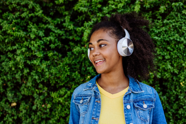 Multiracial girl enjoying music in the headphones, standing in front of green bush, portrait.