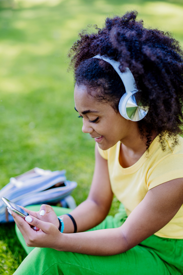 Multiracial girl sitting in a grass and enjoying music in headphones, side view.