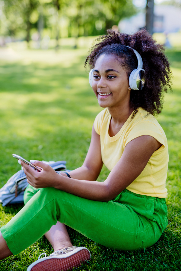 Multiracial girl sitting in a grass and enjoying music in headphones, side view.