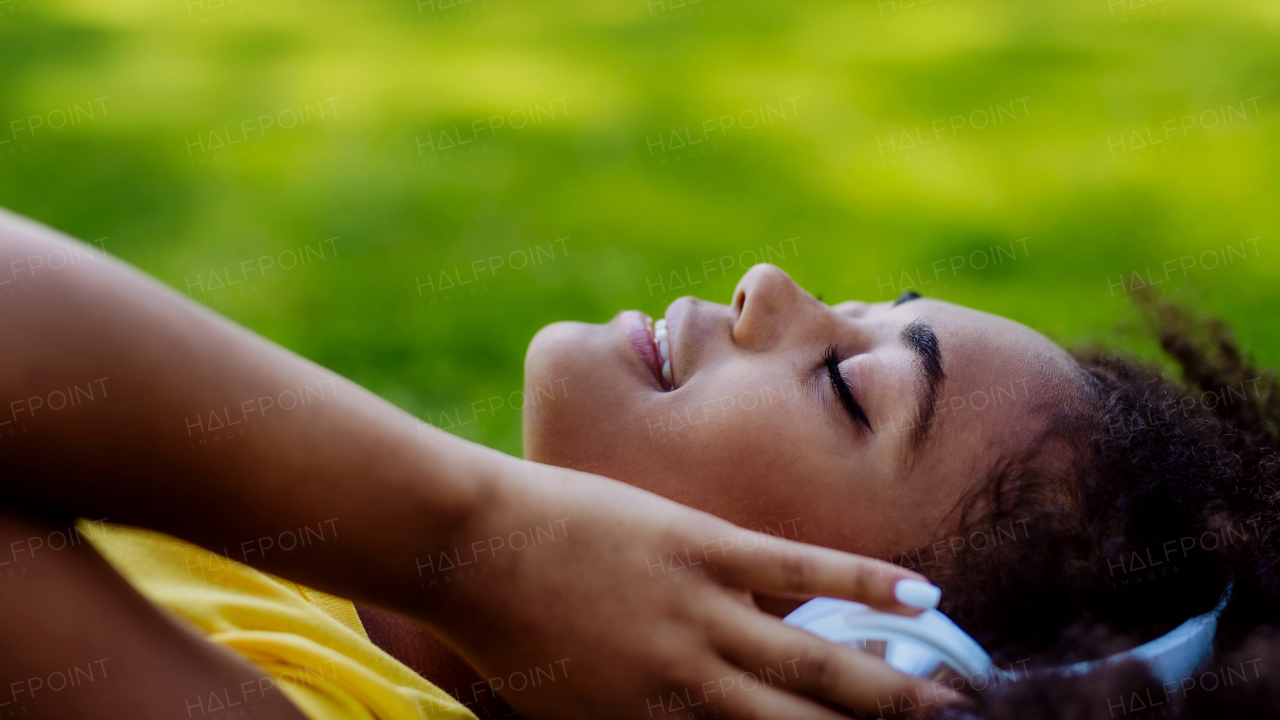 Multiracial girl lying down in a grass and enjoying music in headphones, side view.