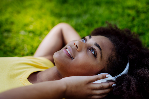 Multiracial girl lying down in a grass and enjoying music in headphones, side view.