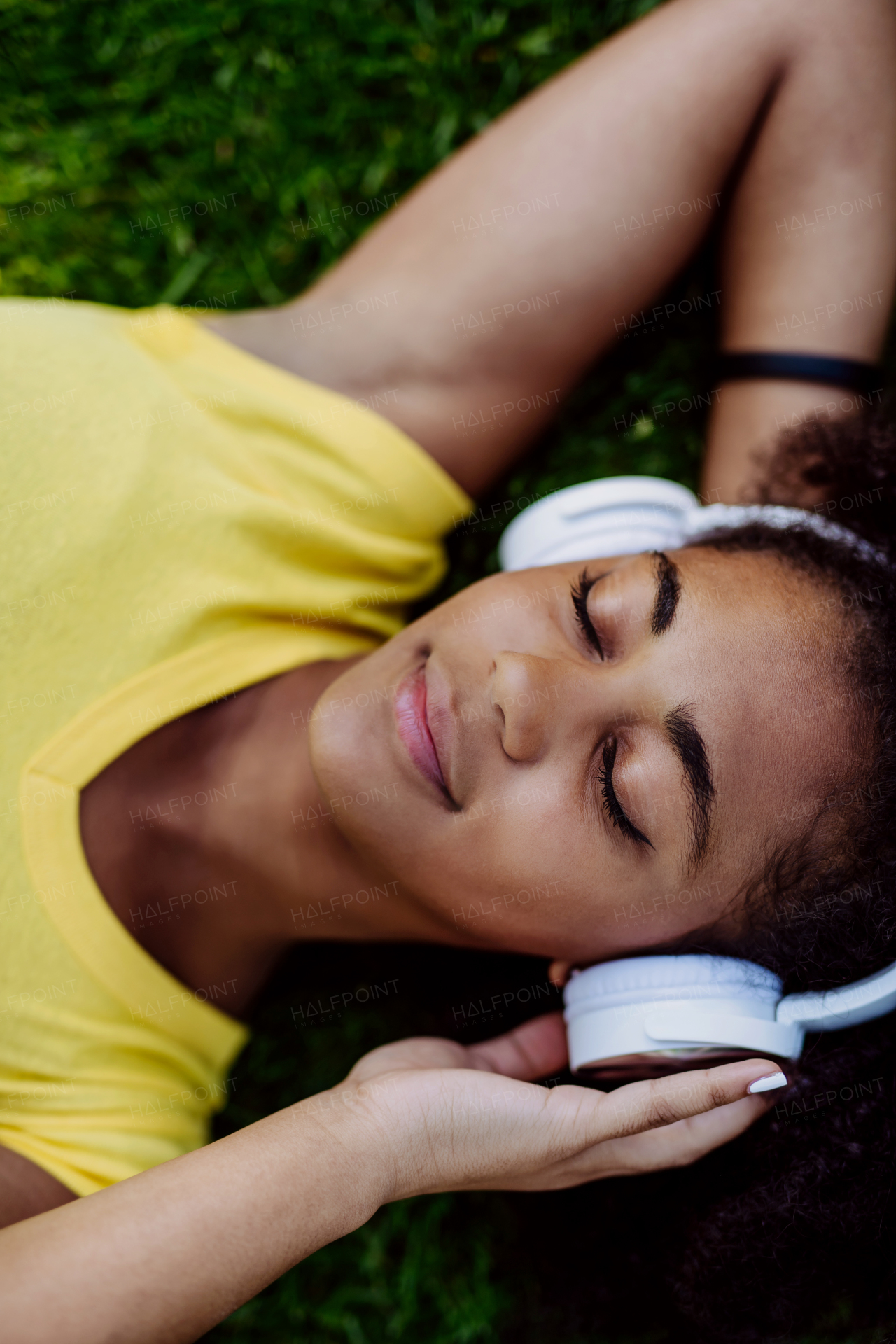 Multiracial girl lying down in a grass and enjoying music in headphones, top view.