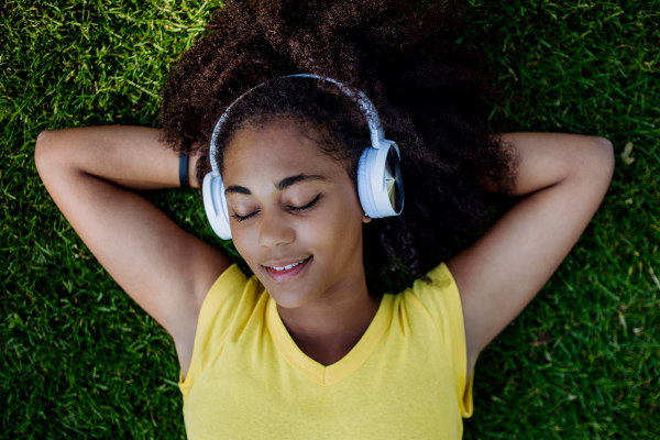 Multiracial girl lying down in a grass and enjoying music in headphones, top view.