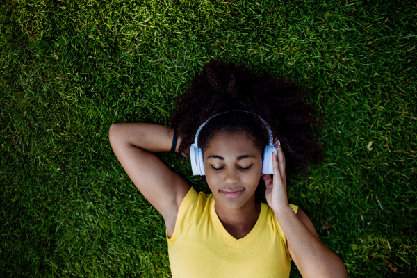 Multiracial girl lying down in a grass and enjoying music in headphones, top view.