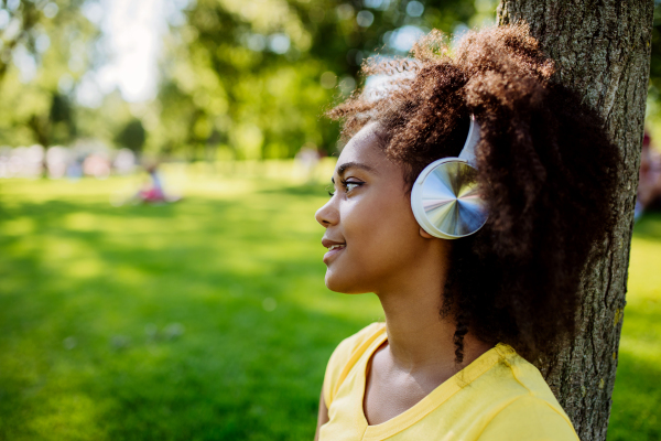 Multiracial girl sitting in a grass and enjoying music in headphones, side view.