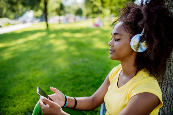 Multiracial girl sitting in a grass and enjoying music in headphones, side view.