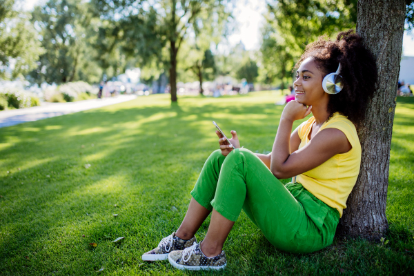 Multiracial girl sitting in a grass and enjoying music in headphones, side view.