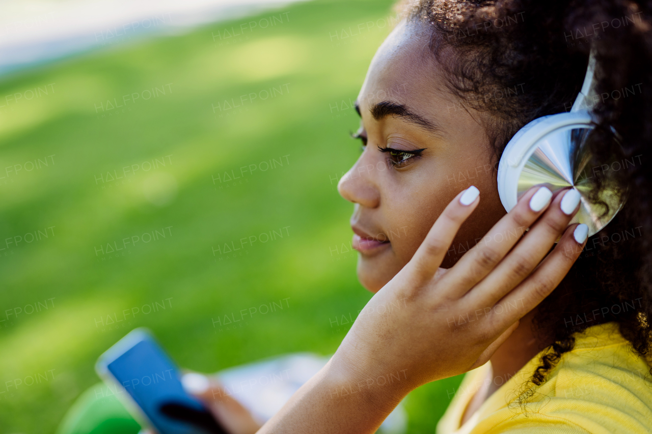 Multiracial girl sitting in a grass and enjoying music in headphones, side view.