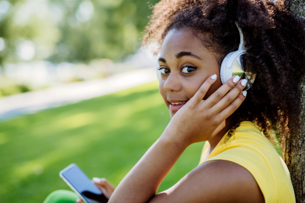 Multiracial girl sitting in a grass and enjoying music in headphones, side view.