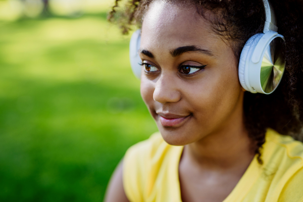 Multiracial girl sitting in a grass and enjoying music in headphones, side view.