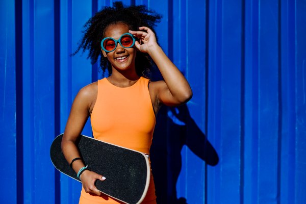 Multiracial teenage girl with skateboard, in front of blue wall during summer day.