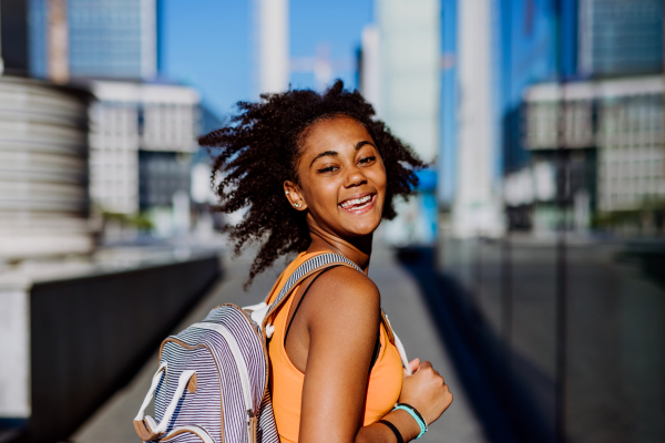 Multiracial teenage girl walking with a backpack in modern city centre during summer day, back to school concept.