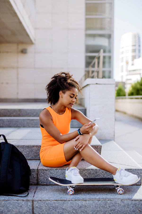 Multiracial teenage girl with backpack and skateboard, sitting and using smartphone in city during a summer day.