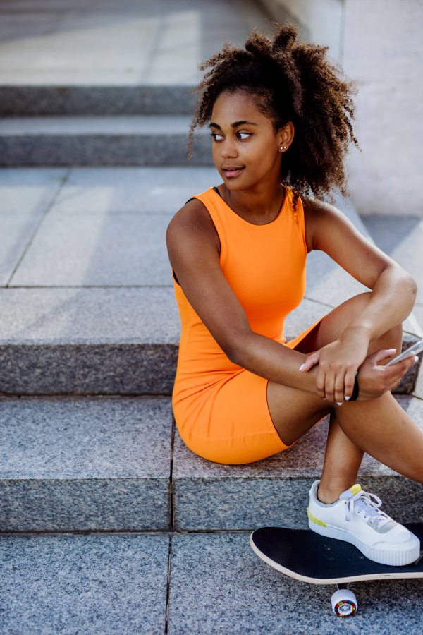 Multiracial teenage girl with backpack and skateboard, sitting and using smartphone in city during a summer day.