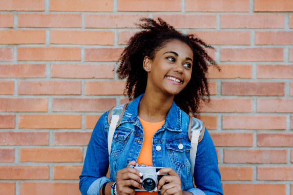 Multiracial girl standing in front of brick wall with a camera, and posing.