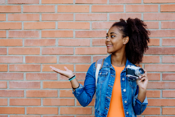 Multiracial girl standing in front of brick wall with a camera, and posing.