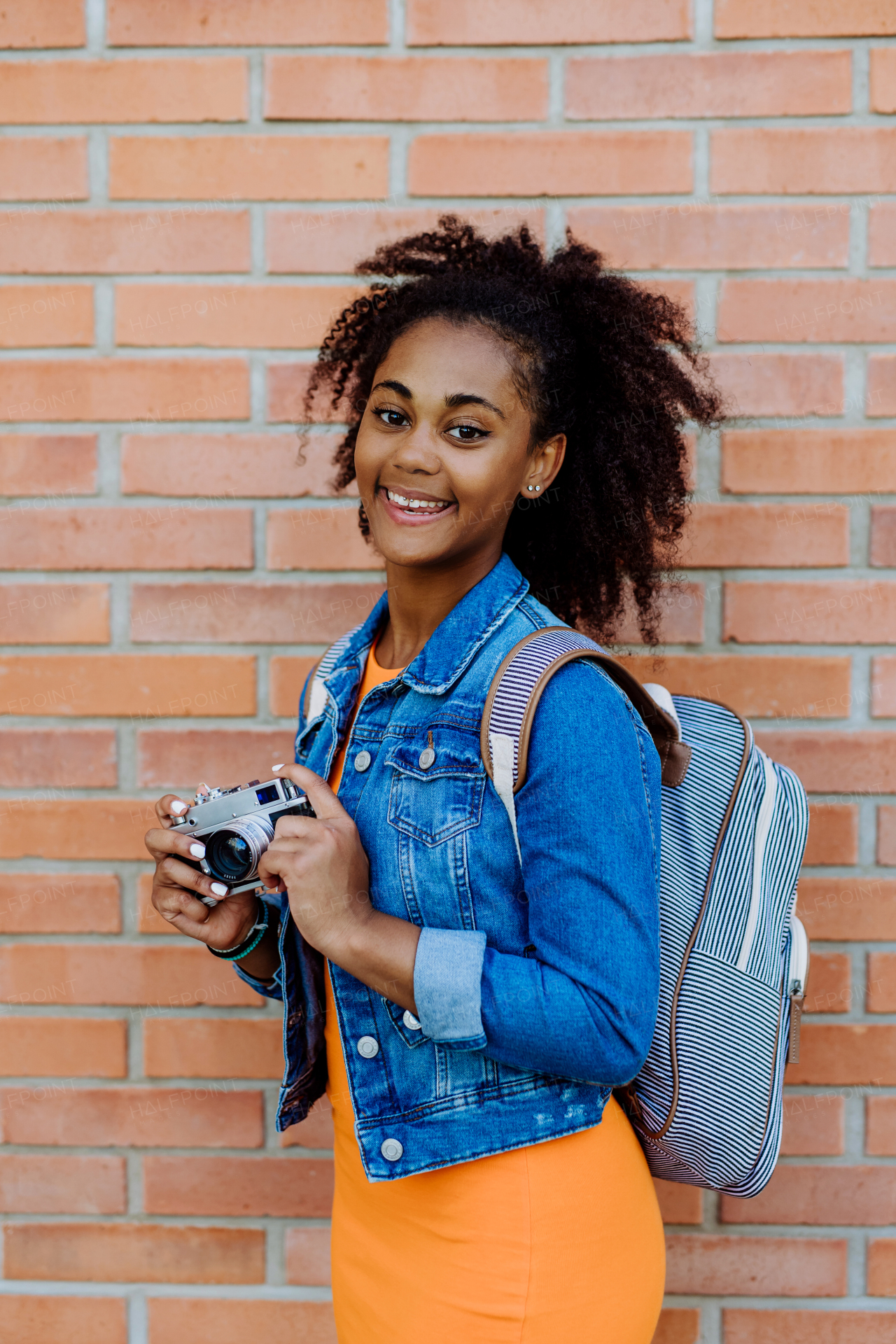 Multiracial girl standing in front of brick wall with a camera, and posing.