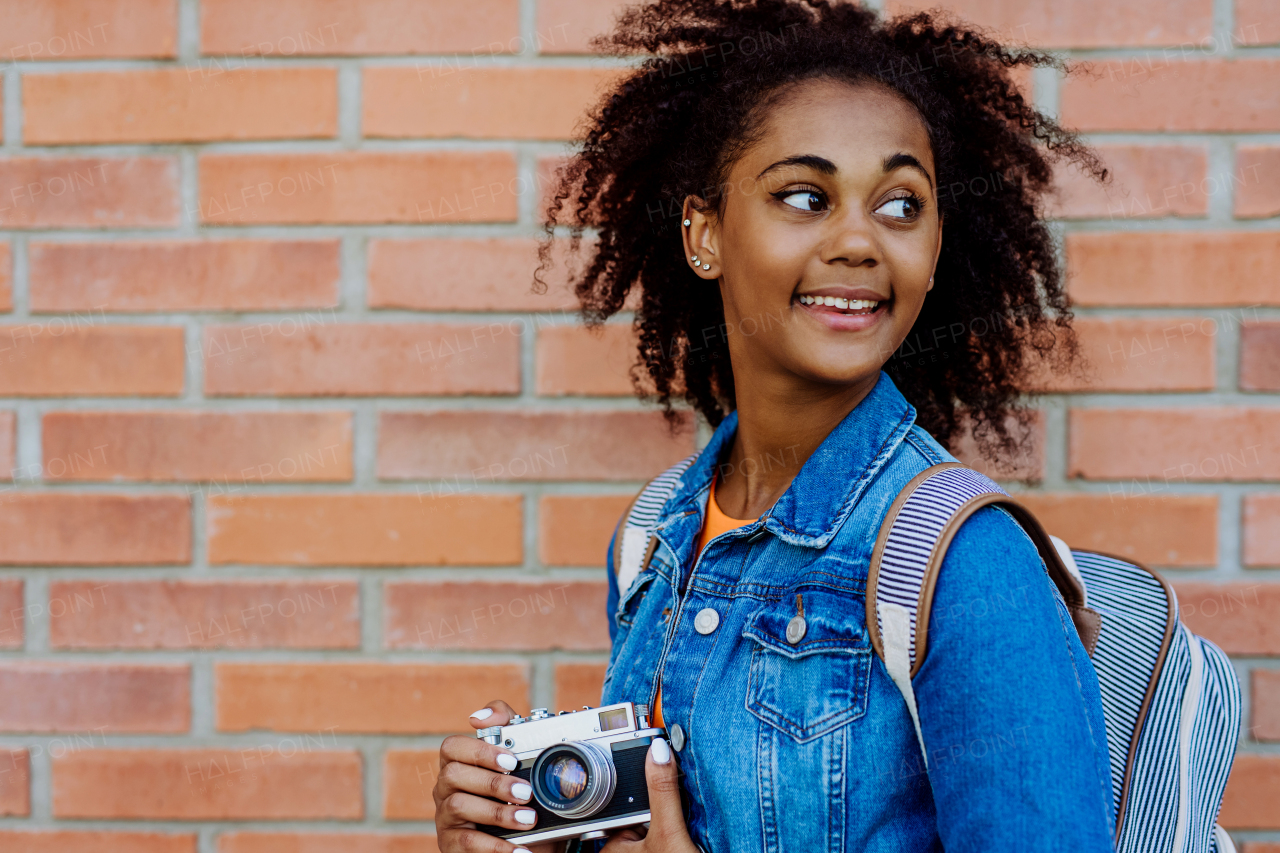 Multiracial girl standing in front of brick wall with a camera, and posing.