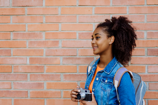 Multiracial teenage girl with a backpack and skateboard, in front of city brick wall.