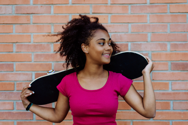 Multiracial teenage girl with backpack and skateboard, walking in city during a summer day.