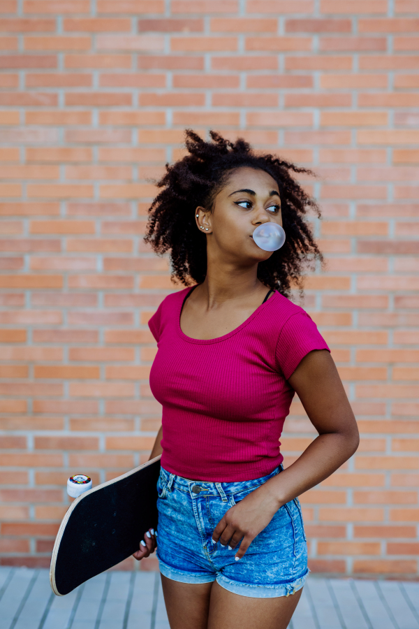 Multiracial teenage girl withchewing gum and skateboard, walking in city during a summer day.