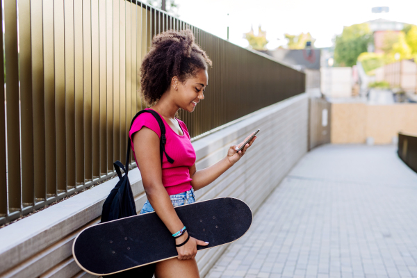 Multiracial teenage girl with backpack and skateboard, walking in city during a summer day.