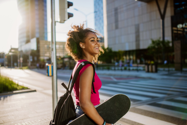 Multiracial teenage girl with backpack and skateboard, walking in city during a summer day.