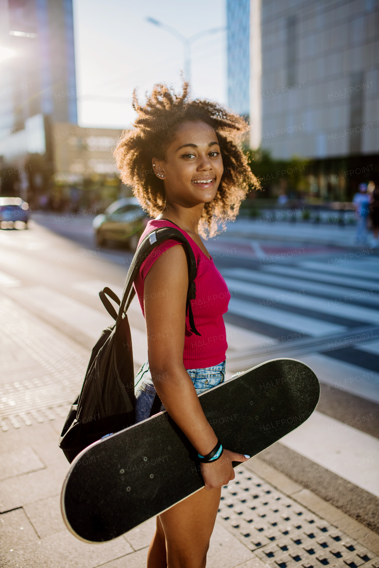 Multiracial teenage girl with backpack and skateboard, walking in city during a summer day.