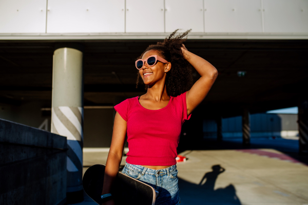 Multiracial teenage girl walking with a skateboard in modern city during summer day.